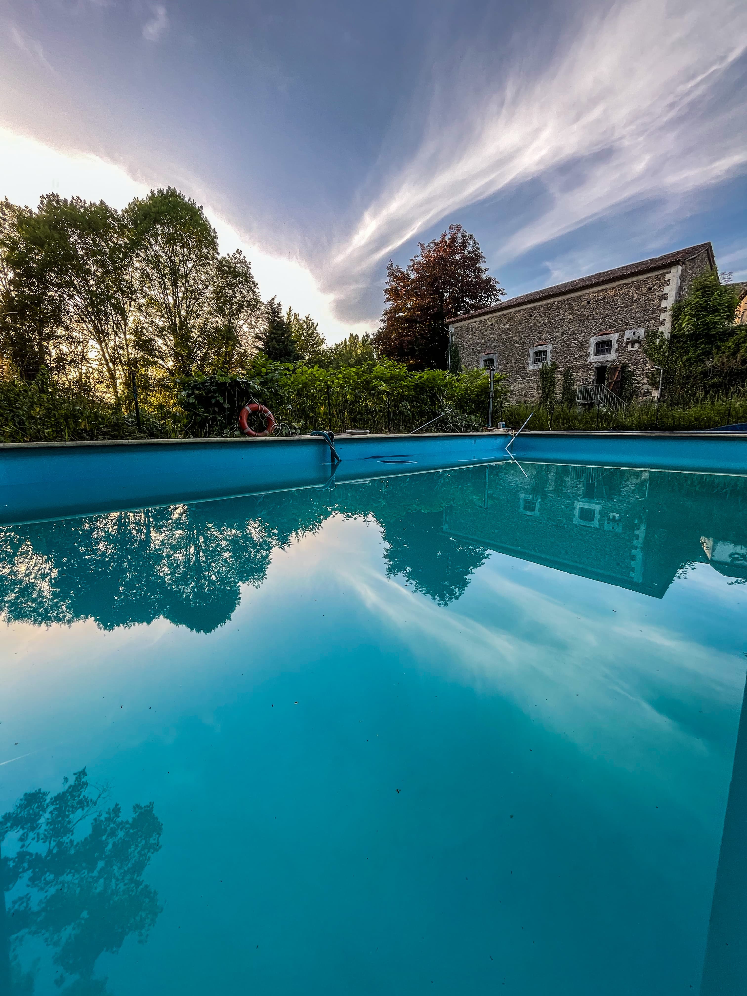 Blue swimming pool reflecting a stone building and trees under a wispy sunset sky.