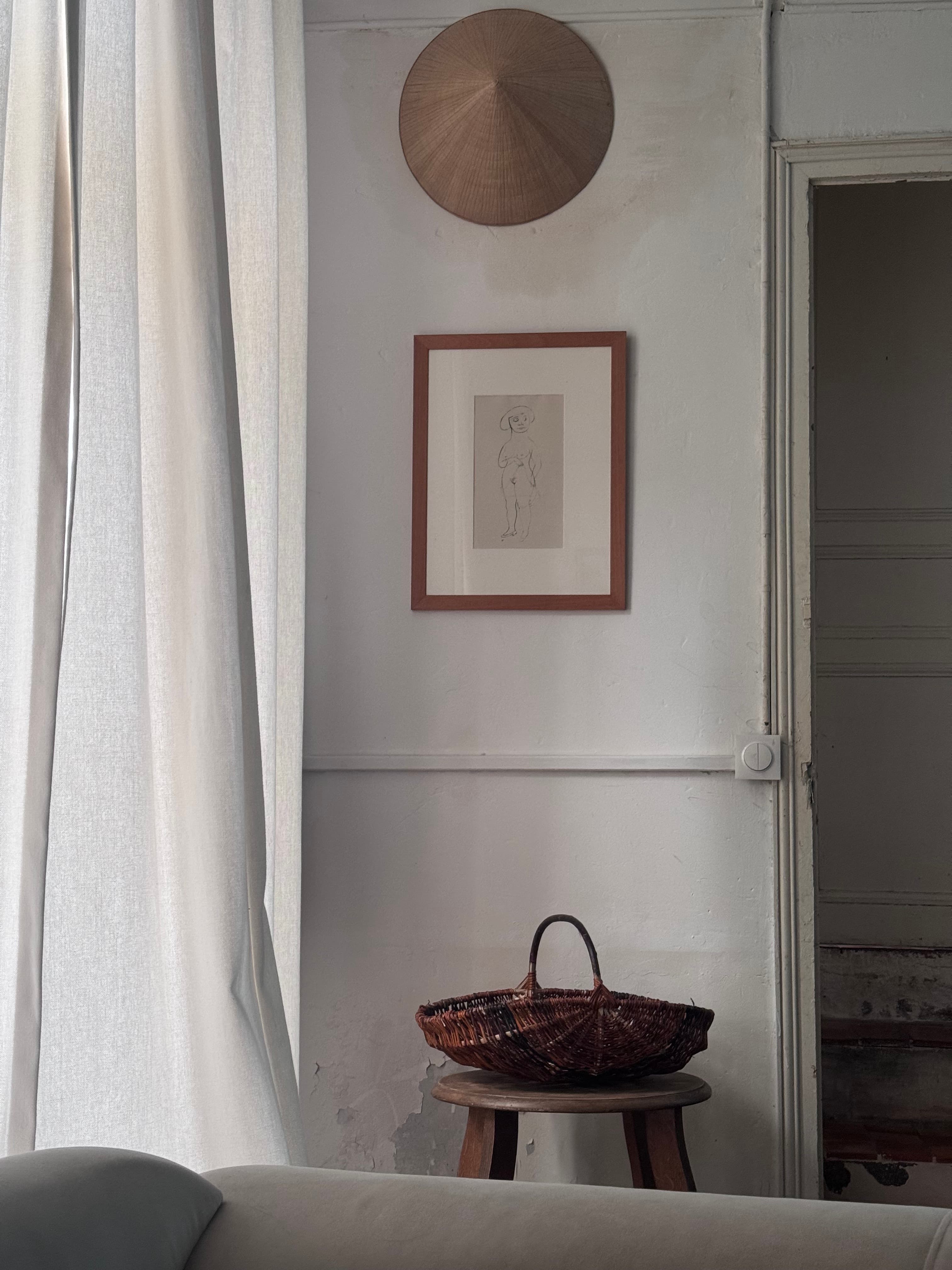 Conical hat, framed sketch, and wicker basket hanging on a white wall above curtains.