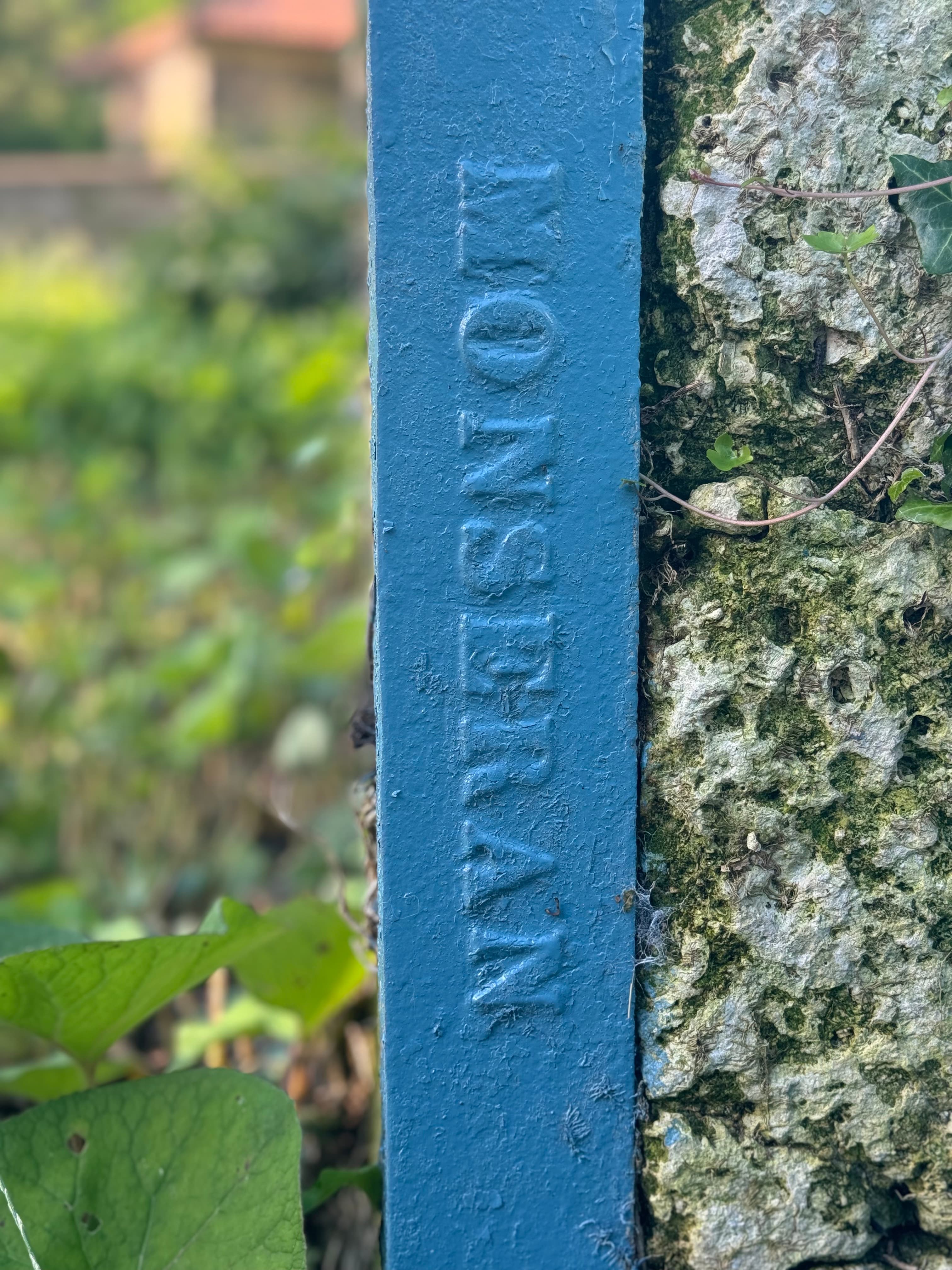 Blue metal post with embossed 'MONSERRAN' text next to a rough, mossy stone wall.