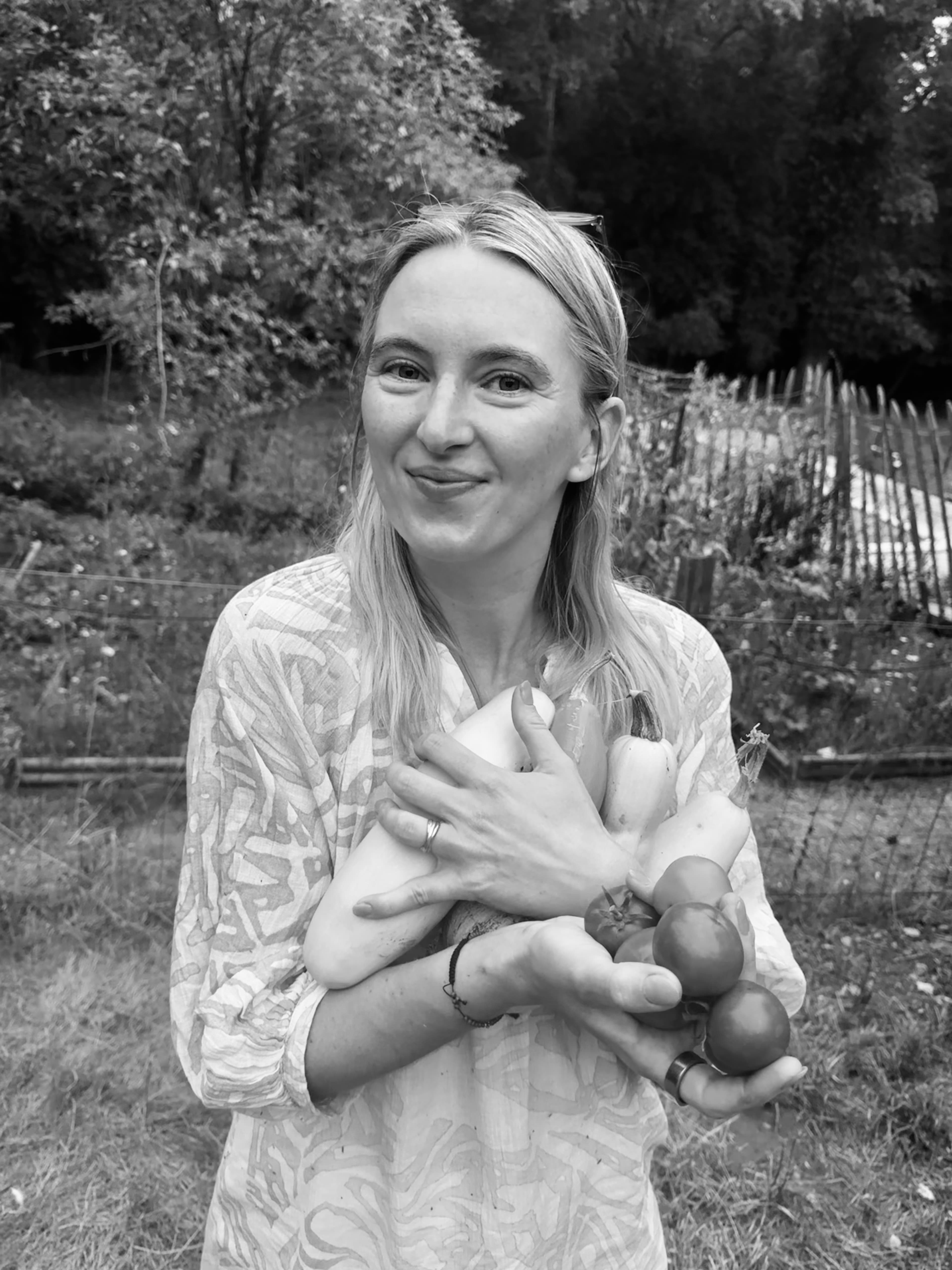 A smiling woman cradles a fresh harvest of squash and tomatoes in her garden.