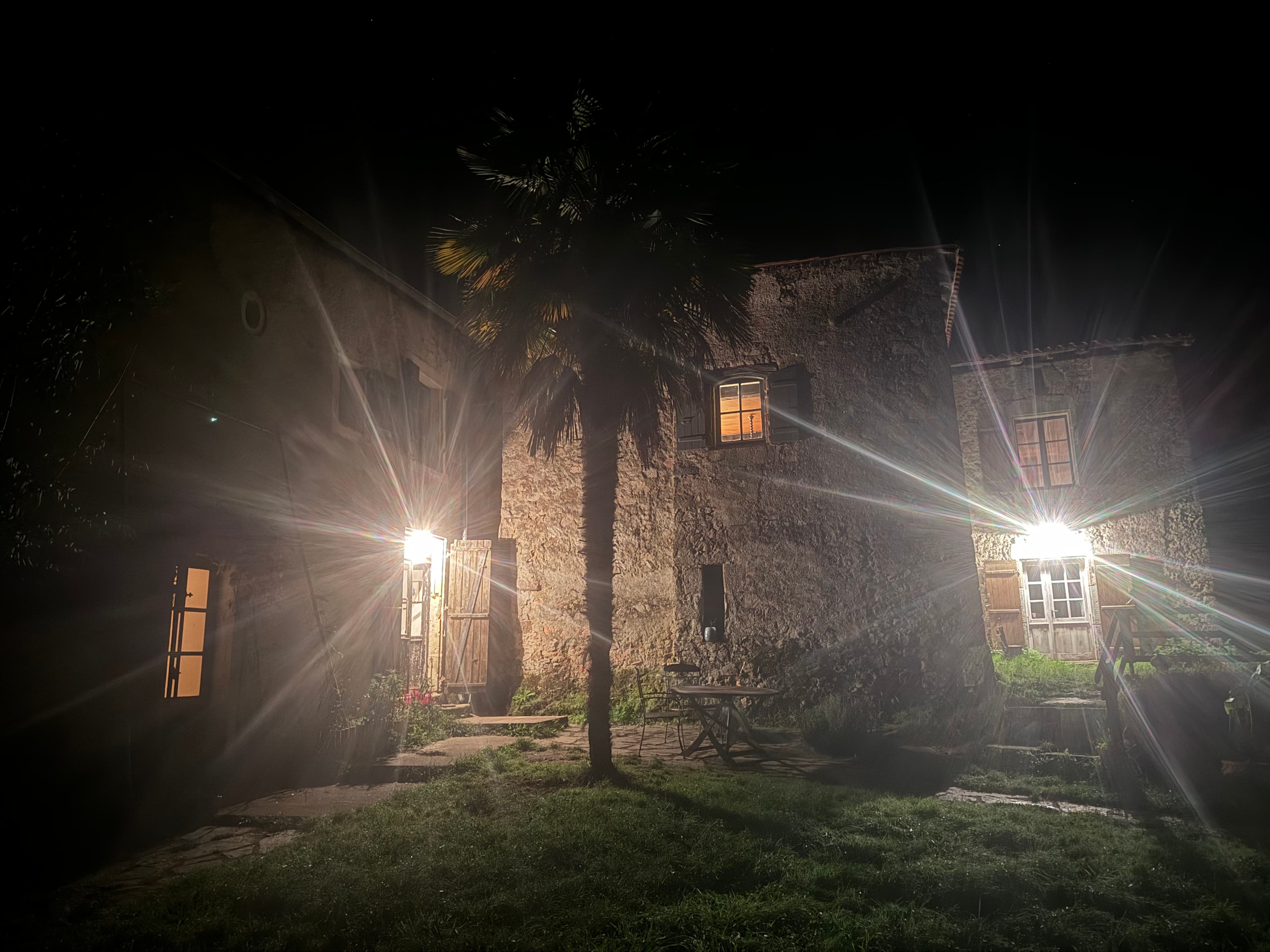 Stone buildings and a palm tree at night, illuminated by bright lights with starburst flares.