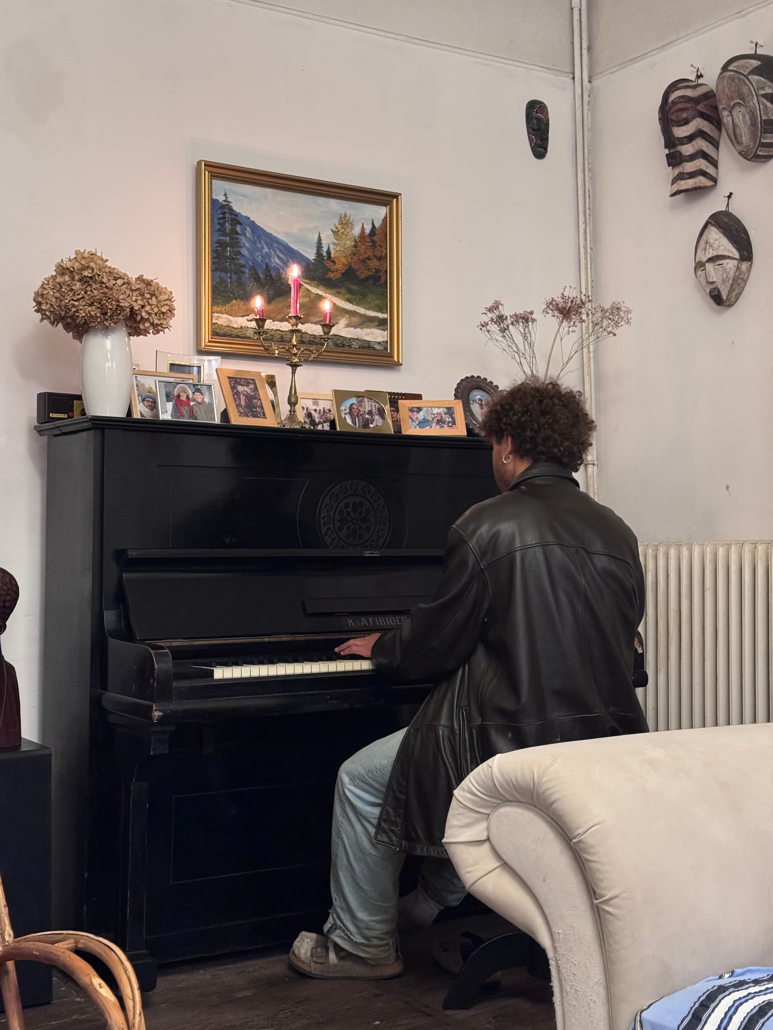Person in a black leather jacket plays a black upright piano in a decorated room.