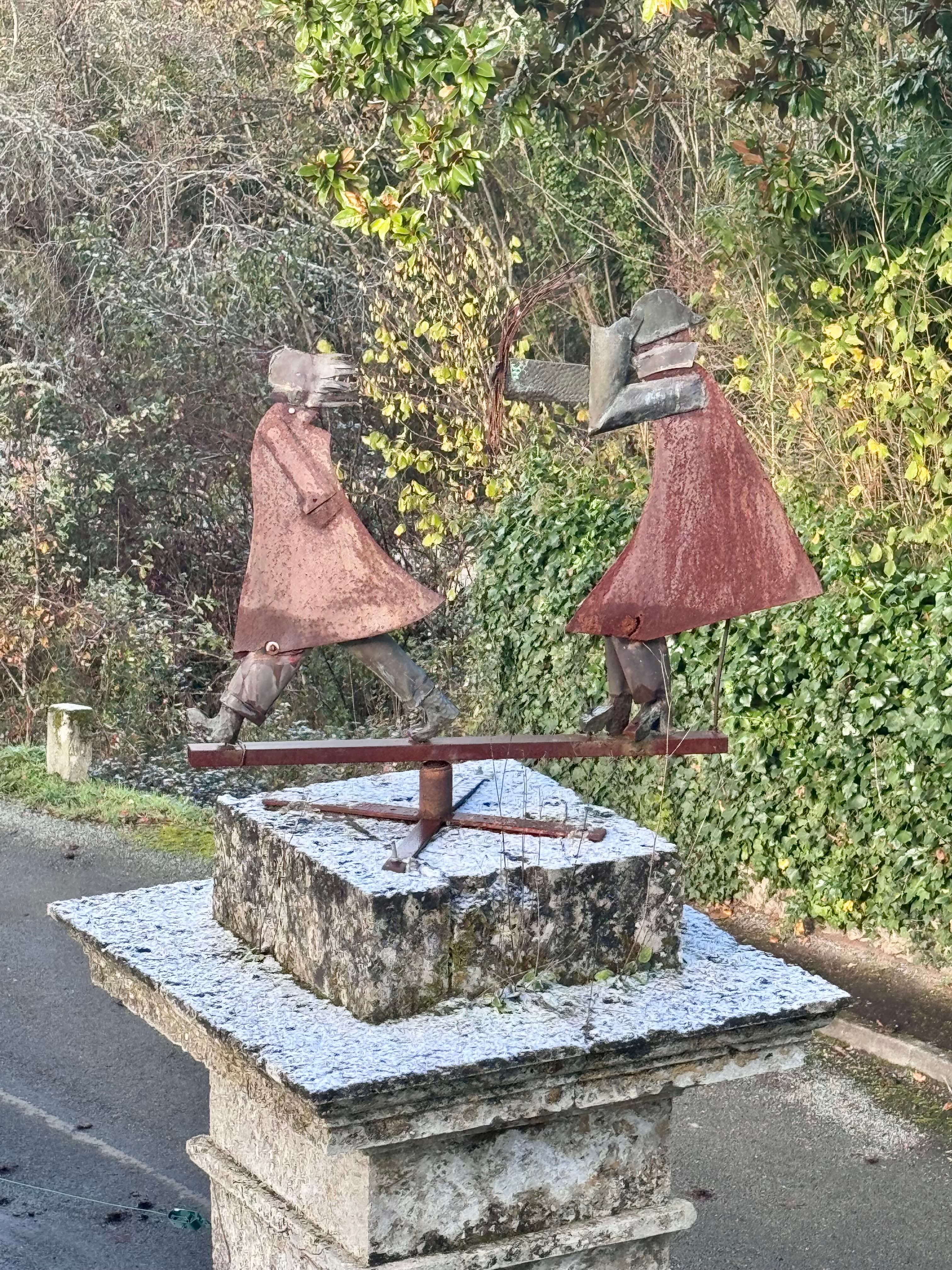 Rusty metal sculpture of two figures on a bar atop a frosted stone pedestal.